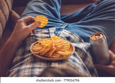After work a guy wearing shirt and jeans lying on sofa, drinking a cold beer, eating crisps and watching sport tv channel. Man's resting time at home concept. 