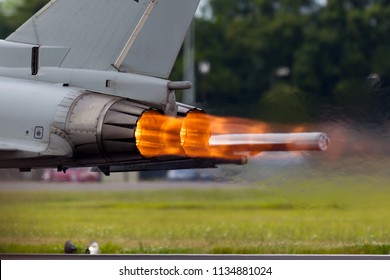 Afterburners glowing on an air force fighter jet aircraft as it speeds down the runway of an air base. 