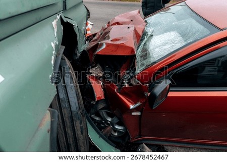 The aftermath of a head-on collision between a car and a truck. The red car is badly damaged: the hood is mangled, the headlight and bumper are broken, and parts are scattered across the road.