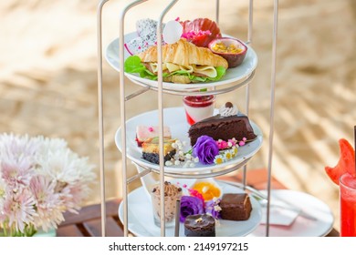 Afternoon tea on tropical sandy beach with shadows from palm trees in hot summer day. Cake stand with fresh pastries, sandwiches, sweeties, shake. Travel in Thailand, luxury hotel and restaurant