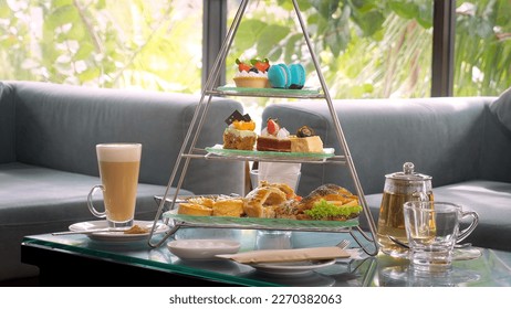 Afternoon tea stand with colorful macarons, petit cakes, and tiny sandwiches, paired with a glass teapot and cup of coffee with tropical palm trees on background. Delicious sweet food.