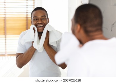 Aftershave Care Concept. Handsome Black Guy Wiping Face With Towel In Bathroom, Young African American Man Looking At Mirror And Smiling While Making Morning Hygiene, Selective Focus On Reflection