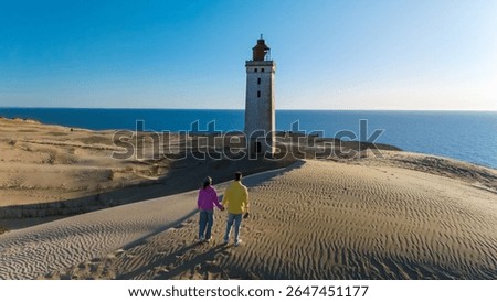 Against a backdrop of pristine dunes and a clear blue sky, a couple strolls hand in hand towards the iconic Rubjerg Knude lighthouse, enjoying a beautiful day by the North Sea of Denmark