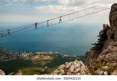 Against the background of the city and the blue sea at a great height, tourists walk over the abyss on rope bridges. Experience the thrill. Mount Aypetri, Yalta, Crimea. - Shutterstock ID 2029432115