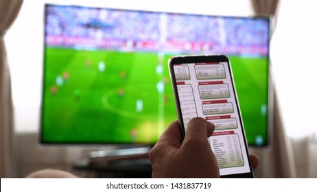 Against The Background Of The TV At Home,  The Guy Orders Different Snacks, Pizza And Sweets Scrolling Through Internet Stores Using Mobile Phones. 