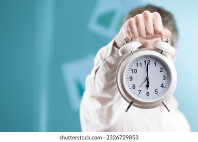 Against a blurry office backdrop, a hand extends a sizable clock. In the corporate world, time is invaluable, and deadlines are near. Stay nimble! - Shutterstock ID 2326729853