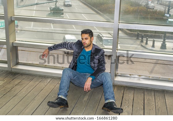 Against a glass wall, a young guy with beard and\
mustache is sitting on the floor into deeply thinking. Background\
is over a street. / Cross\
Road