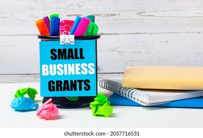 Against A Light Background Of A Wooden Wall, There Is A Glass With Colored Markers, Crumpled Bright Sheets Of Paper And A Sheet Of Blue Paper With The Inscription SMALL BUSINESS GRANTS