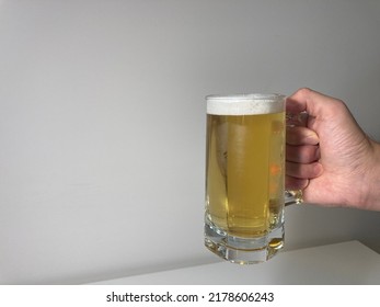 Against A White Background, A Man's Hand Holds A Glass Of Beer. The Beer Is Light And Cold. A Drop Drips Down The Foggy Glass. The Glass Is Full Of Beer, With Foam On Top.