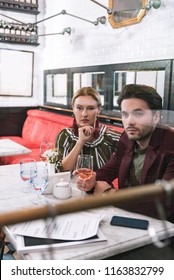 Age Gap. Focused Earnest Woman Touching Face And Man Holding Glass Of Wine