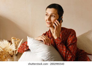 Age, People And Electronic Gadgets Concept. Stylish 50 Year Old Female In Red Shirt Sitting On Comfortable Couch Speaking On Mobile Phone, Talking To Old Friend, Discussing Latest News Or Gossips