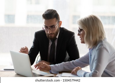 Aged 50s Mentor Female Explain Corporate App To Middle Eastern Ethnicity Employee Use Laptop During Group Meeting In Modern Office, Business People Seated At Desk Negotiating Working On Common Project