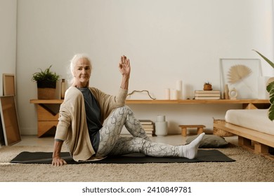 Aged active woman in casualwear sitting on the floor of bedroom and practicing yoga exercises while keeping one hand raised - Powered by Shutterstock