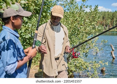 Aged African American Man In Casualwear Holding Rod While Standing In Front Of His Grandson And Fishing Together By Waterside