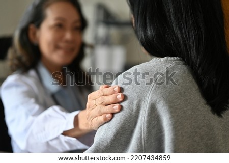 An aged Asian female doctor touching shoulder to comfort and support her patient. A young Asian female patient is being reassured by her doctor. close-up image