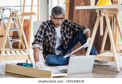 Aged Asian Man With Blueprint Reading Data On Laptop While Making Timber Furniture In Joinery. Older Retired People Hobby And  DIY In Free Time  Concept.