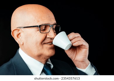 aged bald man is captured up-close, relishing genuine Italian espresso from a suitably-sized cup. Dressed in a dark jacket and white shirt, with black glasses on, he's a picture of contentment. Note: 