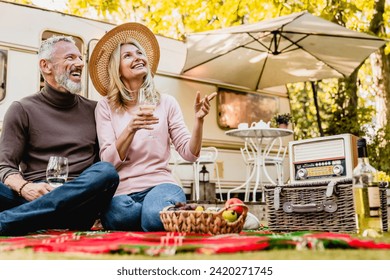 Aged beautiful caucasian couple having picnic and drinking champagne in the yard near the motorhome. Mature blond lady pointing at something - Powered by Shutterstock