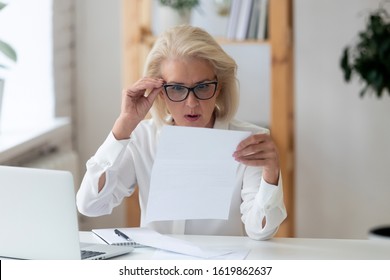 Aged businesswoman sit at workplace desk lowered glasses reads notification feels shocked, bad unexpected news in business letter, financial report, debt notice from bank, bill, taxes increase concept