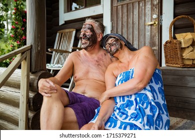 Aged Couple Having Black Facial Mask After Finnish Sauna. Mature Man And Woman In Towels Sitting On Wooden Steps, Relaxing, Laughing And Having Fun. 