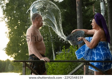 Aged couple having fun after Finnish sauna on wooden cottage pier in a lake. Mature woman pouring cold water from basin over her partner. Typical Finnish summer. 