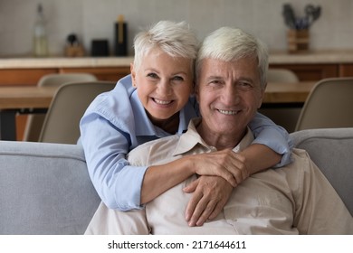 Aged Couple Smile Pose For Camera, Loving Wife Piggyback Husband Seated On Sofa At Home Looking At Camera Laughing Feel Happy. Medical Insurance Cover Ad For Seniors, Homeowner Family Portrait Concept