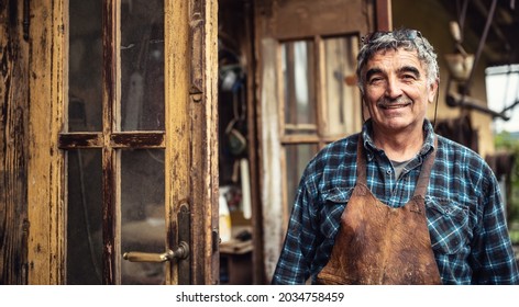Aged craftsman standing and smiling in front of his rustic workshop.