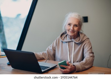 Aged Elderly Woman With Laptop And Mobile Phone Working On Computer At Table