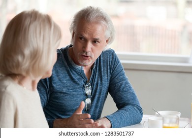 Aged family serious grey haired senior husband telling old wife news having difficulties sitting together at dining room after breakfast, focus on male, spouses communicating spending time together - Powered by Shutterstock