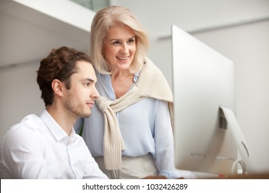 Aged Female Mentor Boss Smiling Looking At Computer Screen Happy For Achievement And Good Online Work Result Of Young Male Intern, Senior Woman Executive Leader Supervising Teaching Helping Colleague