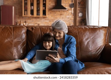 Aged hispanic female babysitter spend time on sofa with preteen girl using tablet pc device watch cartoon play game. Mature granny and little grandchild learn foreign language in online app for kids - Powered by Shutterstock