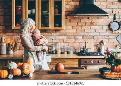 Aged lady with long loose grey hair plays with little granddaughter holding in arms and walking about kitchen against brick wall at home. - Shutterstock ID 1813146328