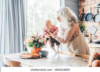 Aged lady with long loose grey hair plays with little granddaughter holding in arms and walking about kitchen against brick wall at home. - Shutterstock ID 1836314833