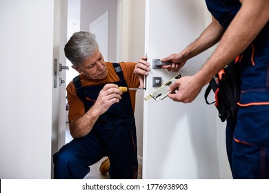 Aged locksmith, repairman, worker in uniform installing, working with house door lock using screwdriver while his colleague bringing him lock plate. Repair, door lock service concept. Focus on man