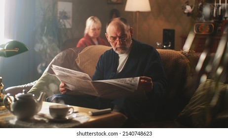 Aged Man In Bathrobe And Glasses Sitting On Couch And Reading Daily Newspaper In Cozy Living Room, Near Woman In Morning At Home