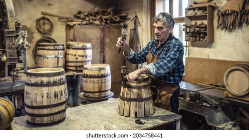 Aged Man Crafts A New Vintage-looking Wooden Barrel Using Hammer To Put The Metal Ring Around The Pieces Of Wood.