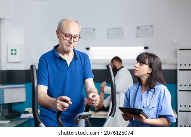 Aged Man Doing Fitness Exercise On Stationary Bicycle For Recovery While Nurse Holding Digital Tablet. Patient With Leg Pain Training For Strength And Healthcare. Adult At Physiotherapy