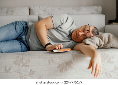 Aged man with glasses fall asleep reading book on comfortable couch in home room. Daytime sleep, napping. Tired old person lying head on pillow on sofa. Stylish soft furniture, relaxation. - Powered by Shutterstock