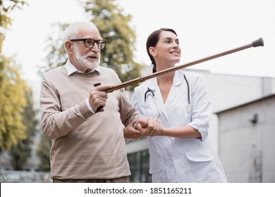 Aged Man Pointing With Walking Stick Away Near Smiling Geriatric Nurse