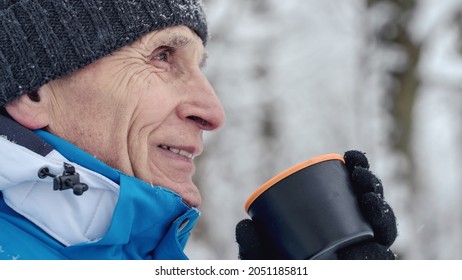 Aged Man In Warm Cap And Gloves Drinks Hot Tea From Thermos Cup In Winter Forest During Hiking Outside