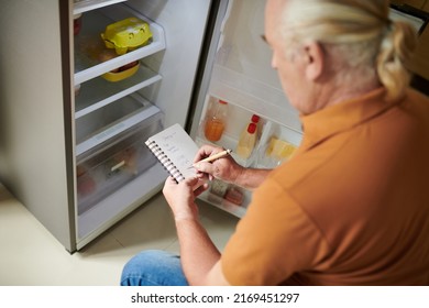 Aged Man Writing Shopping List For Grocery Store When Checking Fridge In Kitchen