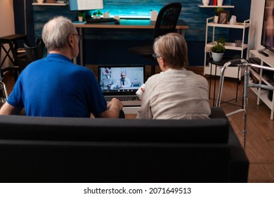 Aged Married Couple Using Telemedicine On Video Call With Laptop In Living Room. Retired Senior People Having Remote Online Checkup Appointment With Doctor While Sitting At Home.