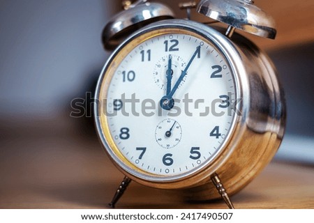 An aged metallic desk alarm clock. It shows noon. It stands out against the blurred background. Idea image about using time efficiently. Selective focus.