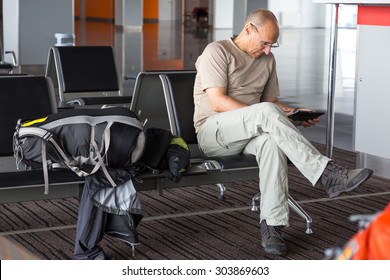 Aged passenger waiting for boarding.
Mature man using tablet PC sitting inside airport terminal building chair line interior day informal simple dress code large cabin luggage sport backpack
