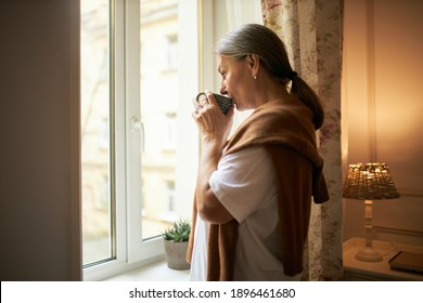 Aged people, lifestyle, retirement and loneliness. Indoor shot of elderly female with gray hair standing by window indoors, spending day at home alone, drinking hot herbal tea, having thoughtful look - Powered by Shutterstock