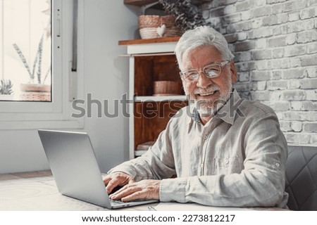 Aged remote worker. Concentrated senior male in glasses work on laptop from home office looking at the camera. Old age man employee freelancer sit at desk table by pc typing report online