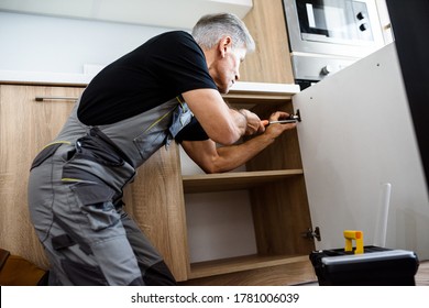 Aged Repairman In Uniform Working, Fixing Kitchen Cabinet Using Screwdriver. Repair Service Concept. Selective Focus On Person. Horizontal Shot. Side View