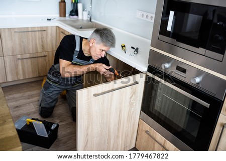 Aged repairman in uniform working, fixing kitchen cabinet using screwdriver. Repair service concept. Selective focus. Horizontal shot. High angle