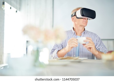 Aged Restired Man With Cup Of Tea Sitting By Table And Watching Virtual Webcast In Vr Goggles