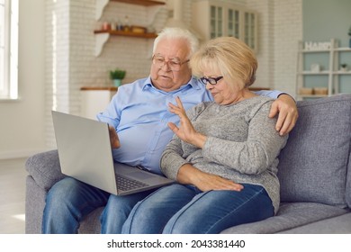 Aged Senior Couple Waving Hands Greeting Grandchildren Through Facetime Using Laptop. Man And Woman Making A Video Call While Sitting At Home On The Couch. Elderly People And Technology Concept.